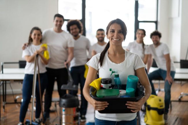 Beautiful cleaning woman with her team at an office holding a bucket with cleaning products all smiling at camera